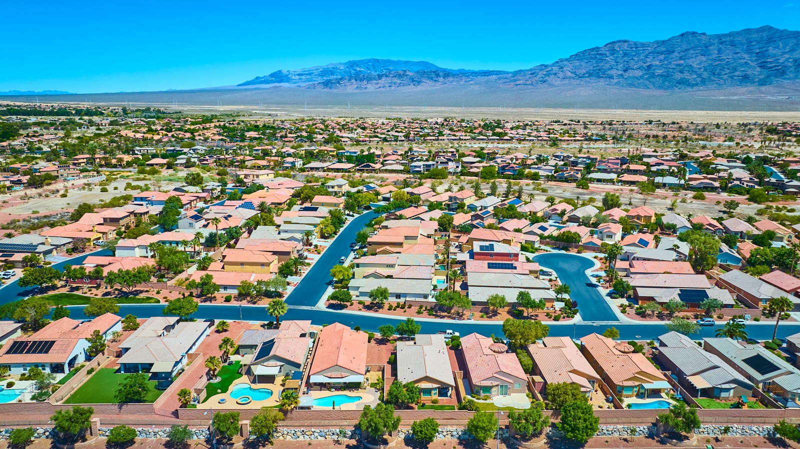 Aerial view of a Las Vegas neighborhood of tiled-roof homes, backyard pools and a canal, with desert mountains behind